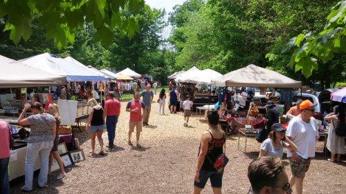 A bustling outdoor market with tents, vendors, and visitors among trees and greenery on a sunny day.