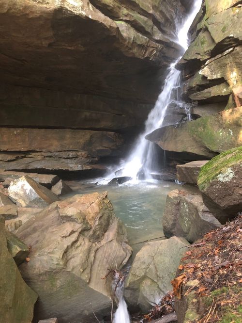 A serene waterfall cascades over rocky cliffs into a clear pool, surrounded by lush greenery and natural stone formations.