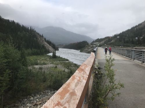 A scenic view of a river winding through a mountainous landscape, with two people walking along a path beside it.