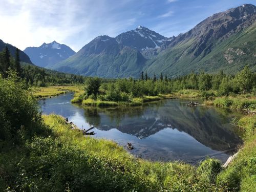 A serene landscape featuring a calm river reflecting mountains and lush greenery under a clear blue sky.