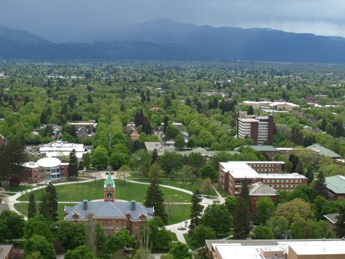 Aerial view of a green campus with buildings, trees, and mountains in the background under a cloudy sky.