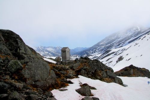 A small wooden structure sits among rocky terrain and snow-covered mountains under a cloudy sky.