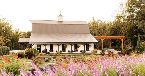 A charming barn-style building surrounded by colorful flowers and greenery, with a wooden pergola nearby.