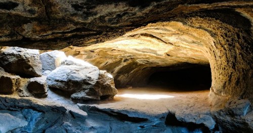 A rocky cave interior with sunlight illuminating the entrance and scattered boulders on the ground.