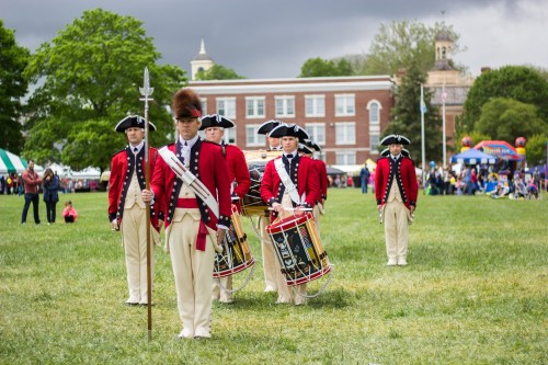 A group of soldiers in red coats and white pants, holding instruments, marching on a grassy field with a crowd in the background.