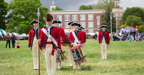 A group of soldiers in red coats and tricorn hats march on a grassy field, with tents and trees in the background.