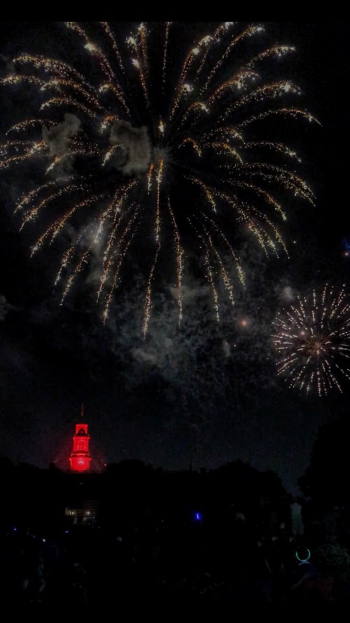 Colorful fireworks illuminate the night sky above a building with a red tower, creating a festive atmosphere.
