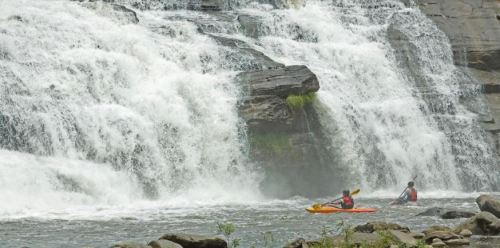 Two kayakers navigate a river near a large waterfall, surrounded by rocky terrain and lush greenery.