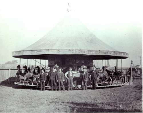 Historic carousel with men standing in front, horses on the ride, and a large tent in the background.