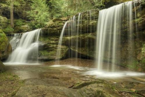 A serene landscape featuring two waterfalls cascading over moss-covered rocks in a lush green forest.