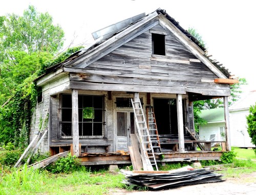 An old, weathered house with a sagging porch, overgrown vegetation, and a ladder leaning against it.