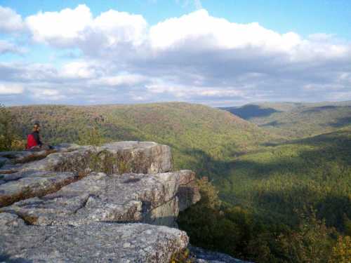 A person sits on a rocky ledge overlooking a lush green valley and rolling hills under a partly cloudy sky.