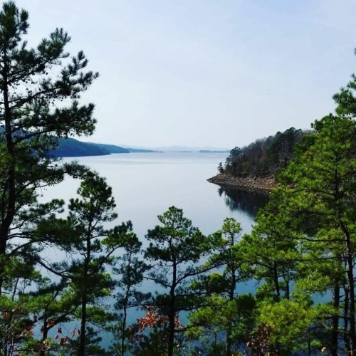 A serene lake surrounded by trees, reflecting the clear sky and distant hills on a calm day.