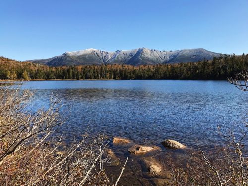 A serene lake surrounded by trees, with mountains in the background under a clear blue sky.