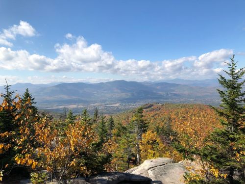 A scenic view of mountains and valleys, with colorful autumn foliage and a clear blue sky dotted with clouds.
