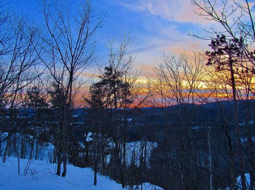 A winter landscape at sunset, featuring bare trees and a colorful sky with shades of orange, pink, and blue.