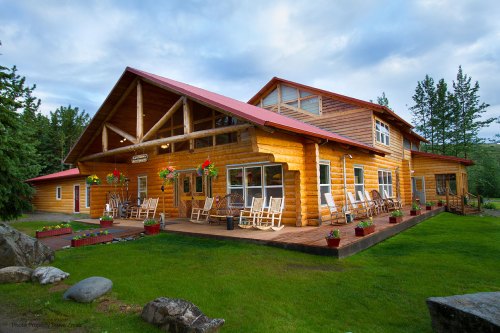 A large wooden lodge with a red roof, surrounded by greenery and rocking chairs on the porch, under a cloudy sky.