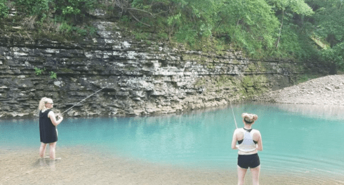 Two people fishing in a clear blue river surrounded by rocky cliffs and lush greenery.