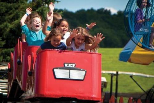 A group of children joyfully riding a roller coaster, hands raised in excitement against a sunny backdrop.