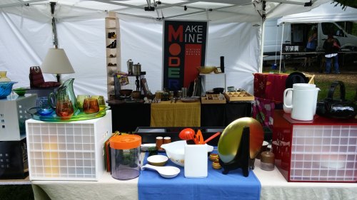 A colorful market stall displaying various modern kitchenware and decorative items under a white tent.