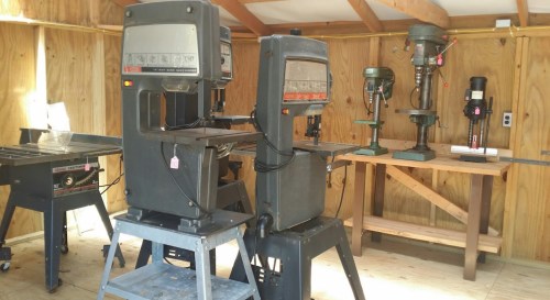 A workshop with various woodworking machines, including band saws and drill presses, set against wooden walls.