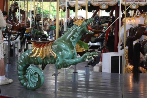 A colorful carousel figure of a dragon with wings, set against a backdrop of other carousel animals.