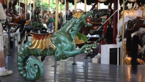 A colorful carousel figure of a dragon-like creature with wings, set against a blurred background of other carousel animals.
