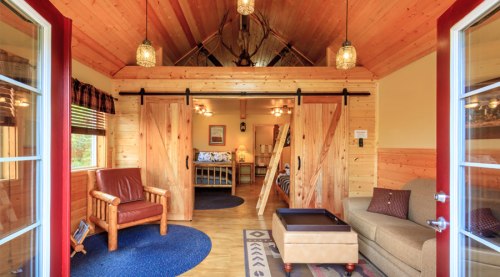 Cozy wooden interior with sliding barn doors, a leather chair, sofa, and warm lighting in a rustic cabin setting.