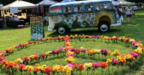 A colorful peace sign made of flowers in front of a decorated van at a festival, with tents in the background.
