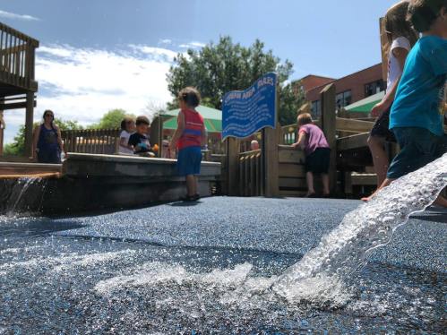 Children play at a water feature in a playground, with a sign and trees in the background under a sunny sky.