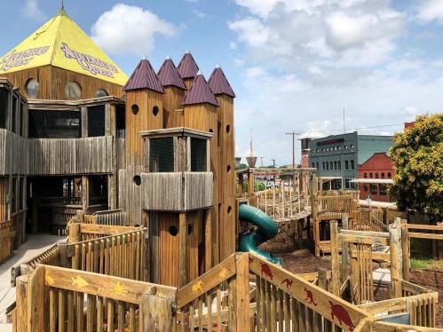 A wooden playground with a castle-like structure, slides, and climbing areas, set against a blue sky.
