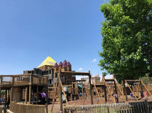 A large wooden playground with slides and swings, surrounded by trees under a clear blue sky.