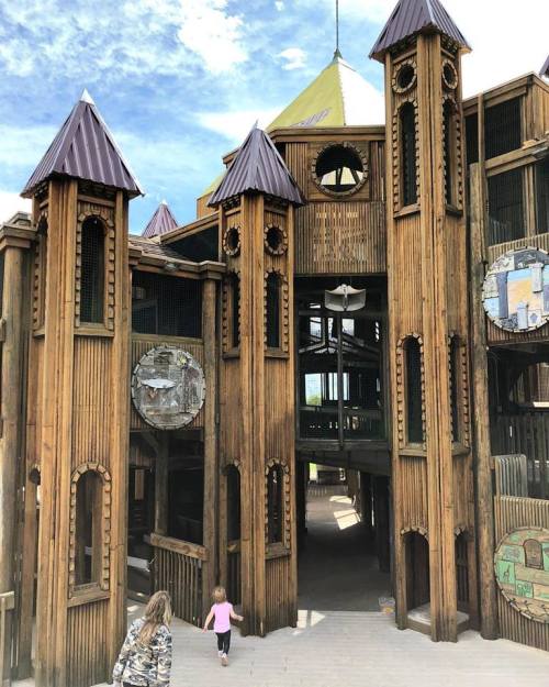 Children exploring a large wooden playground structure with towers and a colorful design under a blue sky.