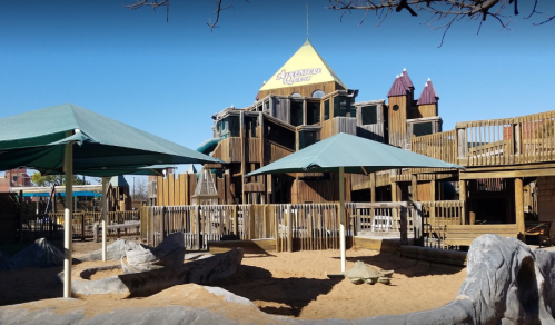 A large wooden playground with towers and slides, surrounded by sandy areas and shaded by green canopies.