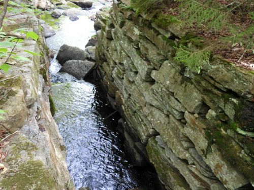 A narrow waterway flows between moss-covered stone walls, surrounded by greenery and rocks.