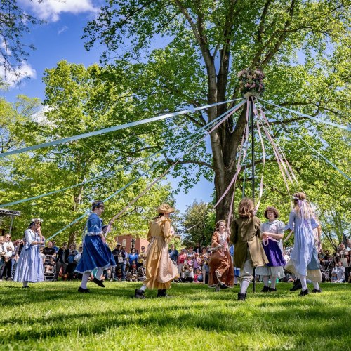 A group of children dances around a maypole, adorned with ribbons, under a sunny sky and surrounded by spectators.