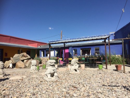Colorful buildings surround a gravel courtyard with stone sculptures and potted plants under a clear blue sky.
