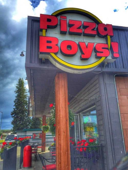 Sign for "Pizza Boys!" on a building, with red flowers and outdoor seating under a cloudy sky.