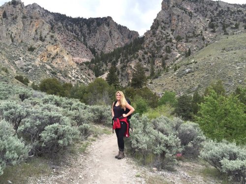 A woman stands on a trail surrounded by green shrubs and rocky mountains in the background.