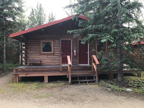 A rustic log cabin with a red roof, surrounded by trees, featuring a porch and two front doors.