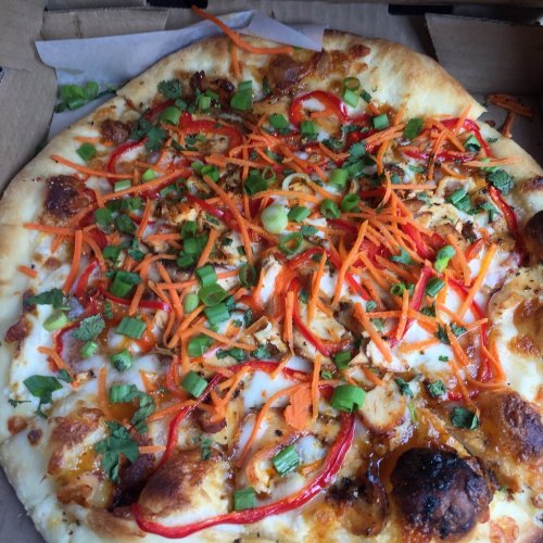 A pizza topped with shredded carrots, red peppers, green onions, and herbs on a golden crust in a pizza box.