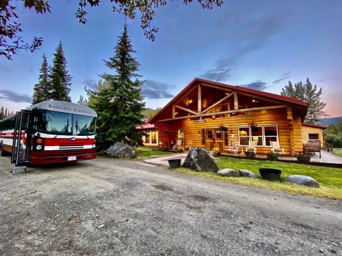 A rustic log cabin with a red roof, surrounded by trees, and a bus parked nearby on a gravel path at sunset.