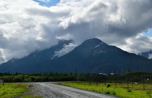 A gravel road leads to majestic mountains, partially covered by clouds, with greenery in the foreground.