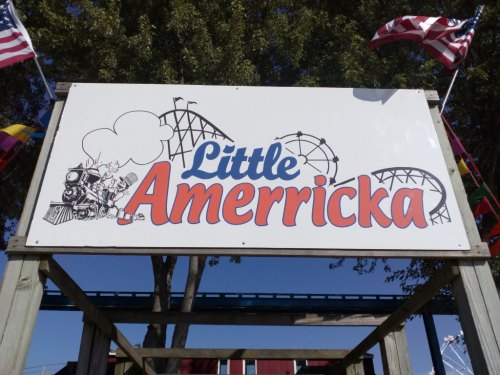 Sign for "Little Americka" amusement park featuring roller coasters and colorful flags against a clear blue sky.