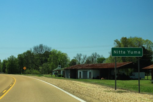 Roadside view of a sign for Nitta Yuma, with green trees and a rustic building in the background. Clear blue sky.