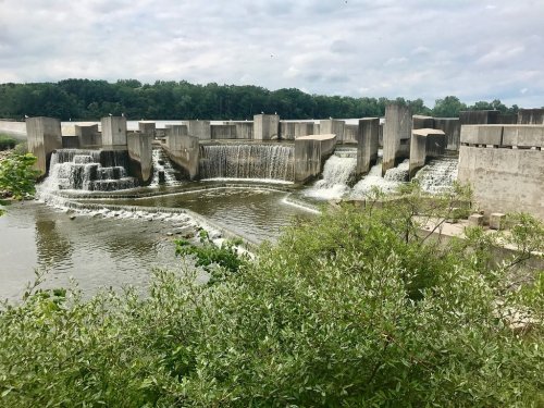 A concrete dam with cascading water, surrounded by greenery and a cloudy sky in the background.