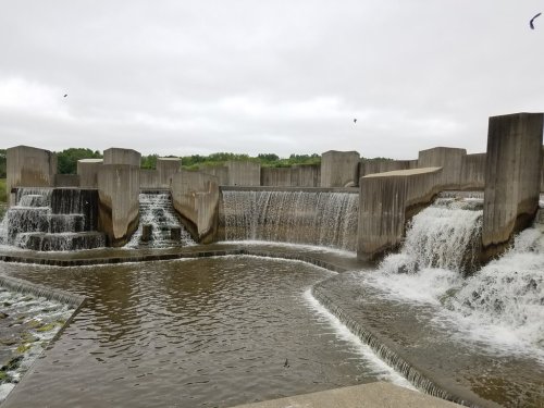 A concrete structure with cascading waterfalls flows into a calm body of water under a cloudy sky.