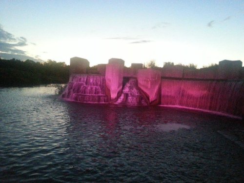 A waterfall cascading over a dam, illuminated in pink light at dusk, with a calm water surface reflecting the colors.