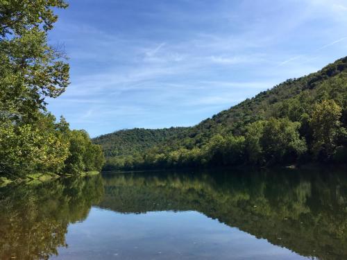 A serene river reflects lush green hills under a clear blue sky, surrounded by trees.
