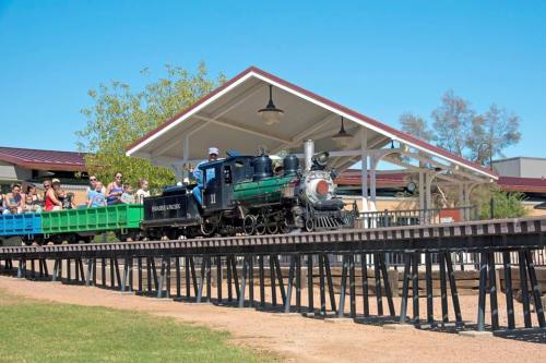 A miniature train on a raised track, with passengers enjoying a ride under a shaded pavilion on a sunny day.